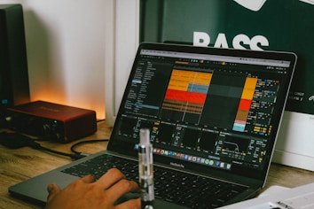A person is using a MacBook Pro laptop with music production software open on the screen, displaying a project with multiple tracks and controls. The laptop is placed on a wooden desk beside a red audio interface. In the background, part of a poster with the word 'BASS' is visible.