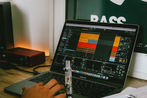 A person is using a MacBook Pro laptop with music production software open on the screen, displaying a project with multiple tracks and controls. The laptop is placed on a wooden desk beside a red audio interface. In the background, part of a poster with the word 'BASS' is visible.