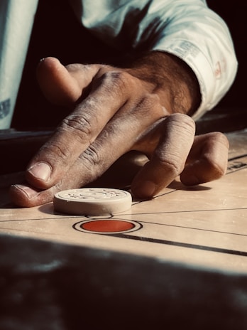 An overhead shot of a carrom board mid-game, with hands poised to strike the striker.