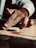 A hand is poised on a carrom board, finger lined up to flick a white carrom disc towards a red circle target. The background shows a blurred view of the board's surface, with playworn textures.