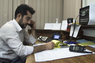 A man with a beard and short hair is seated at a wooden desk, holding a phone to his ear while writing on papers in a notepad. The setting appears to be an office environment with various items on the desk including a telephone, a radio communication device, a computer monitor, and numerous sheets of paper and documents.