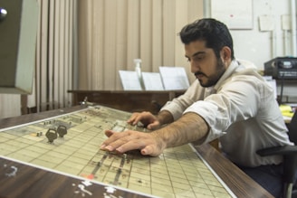 A man is concentrating as he operates a vintage control panel in an office environment. He is using both hands to adjust the settings on the panel, suggesting focus and careful attention. The room contains office equipment and papers are visible in the background.