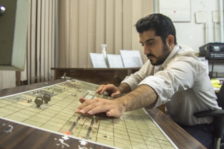 A man is concentrating as he operates a vintage control panel in an office environment. He is using both hands to adjust the settings on the panel, suggesting focus and careful attention. The room contains office equipment and papers are visible in the background.