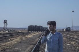 A man is standing on a railway track holding a walkie-talkie to his mouth. He is wearing a blue shirt and appears to be communicating through the device. Behind him are several train cars on the tracks, and a distant landscape with sparse vegetation and a distant watchtower on the left.
