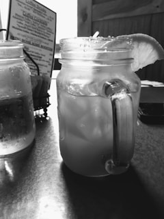 A mason jar filled with a cold beverage, possibly lemonade, with ice cubes and a lemon slice on the rim. The jar is sitting on a table with another jar partially visible next to it. In the background, there is a menu and a wooden wall.