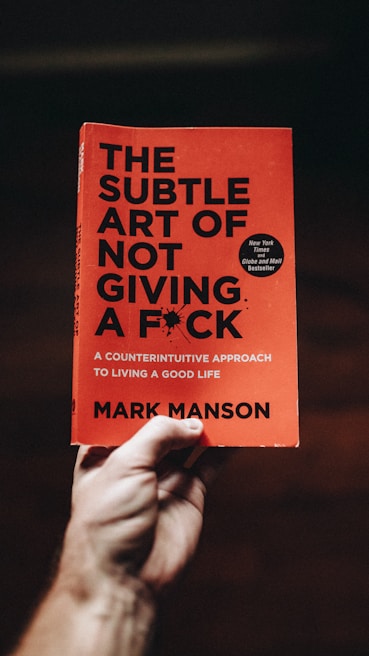 A hand holds a book titled 'The Subtle Art of Not Giving a F*ck' by Mark Manson. The cover is orange with black text, featuring a small black burst graphic and a circular sticker indicating it is a bestseller.