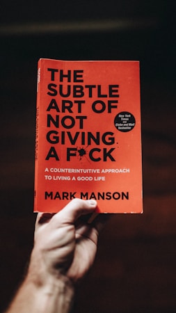 A hand holds a book titled 'The Subtle Art of Not Giving a F*ck' by Mark Manson. The cover is orange with black text, featuring a small black burst graphic and a circular sticker indicating it is a bestseller.