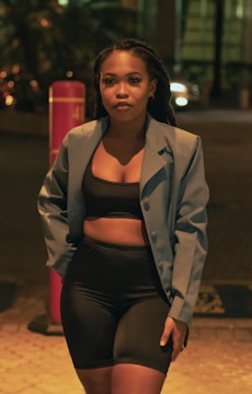 A woman stands in an outdoor setting at night. She is wearing a light blue blazer over a black crop top and black shorts. The background shows some blurred objects and lights, possibly from a street or urban environment.
