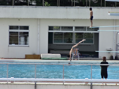 A diving pool scene with a person on a diving board preparing to dive and another person executing a handstand near the poolside. The setting includes a building with large windows in the background and several individuals in swimwear, indicating an environment of training or recreation.