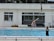 A diving pool scene with a person on a diving board preparing to dive and another person executing a handstand near the poolside. The setting includes a building with large windows in the background and several individuals in swimwear, indicating an environment of training or recreation.