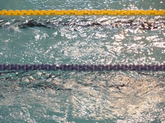 Choppy water in a swimming pool with two parallel float lines, one yellow and one blue. The water is sparkling under bright lighting, creating reflections and shadows.