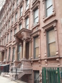 A row of classic brownstone buildings with ornate architectural details, including large windows and decorative columns. The entrance features a set of steps leading up to a covered porch area. Air conditioning units are visible in several windows along the facade.