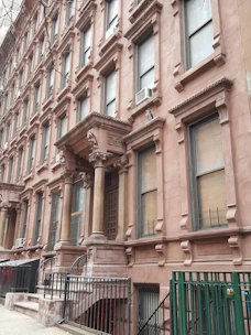 A row of classic brownstone buildings with ornate architectural details, including large windows and decorative columns. The entrance features a set of steps leading up to a covered porch area. Air conditioning units are visible in several windows along the facade.