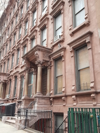 A row of classic brownstone buildings with ornate architectural details, including large windows and decorative columns. The entrance features a set of steps leading up to a covered porch area. Air conditioning units are visible in several windows along the facade.