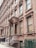A row of classic brownstone buildings with ornate architectural details, including large windows and decorative columns. The entrance features a set of steps leading up to a covered porch area. Air conditioning units are visible in several windows along the facade.