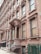 A row of classic brownstone buildings with ornate architectural details, including large windows and decorative columns. The entrance features a set of steps leading up to a covered porch area. Air conditioning units are visible in several windows along the facade.