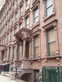 A row of classic brownstone buildings with ornate architectural details, including large windows and decorative columns. The entrance features a set of steps leading up to a covered porch area. Air conditioning units are visible in several windows along the facade.