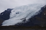 A massive glacier cascades down a dark rocky mountainside with intricate patterns of ice and snow. The sky is overcast, lending a moody atmosphere.