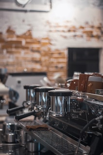 A commercial espresso machine with multiple portafilters and a row of cups on top, set against a background of a rustic, textured wall with a mix of exposed bricks and plaster.