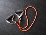 An orange resistance band with grey foam handles is placed on a textured black surface. The handles have red stripes, and the band is coiled in a loop shape.