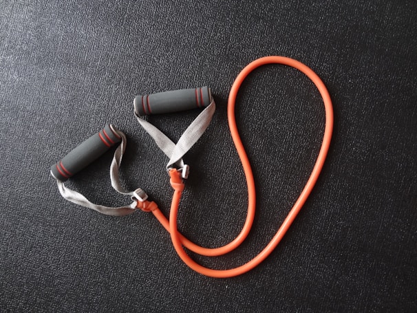 An orange resistance band with grey foam handles is placed on a textured black surface. The handles have red stripes, and the band is coiled in a loop shape.