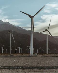 Several large wind turbines stand against a backdrop of mountains and cloudy skies, positioned on a barren desert landscape. The scene conveys a sense of isolation and renewable energy.