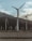 Several large wind turbines stand against a backdrop of mountains and cloudy skies, positioned on a barren desert landscape. The scene conveys a sense of isolation and renewable energy.
