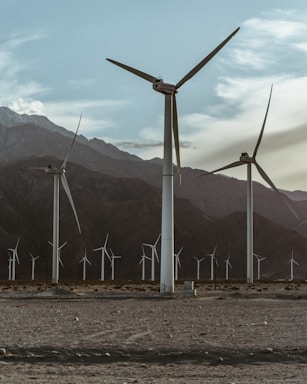 Several large wind turbines stand against a backdrop of mountains and cloudy skies, positioned on a barren desert landscape. The scene conveys a sense of isolation and renewable energy.