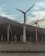 Several large wind turbines stand against a backdrop of mountains and cloudy skies, positioned on a barren desert landscape. The scene conveys a sense of isolation and renewable energy.