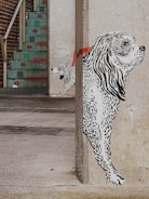 A mural of two black and white dogs is painted on the corner of a concrete wall. The larger dog appears to be in the foreground, with detailed line work illustrating its fur. The smaller dog is positioned behind it, partially hidden. A red stripe extends across the wall behind the dogs. In the background, there is a staircase with turquoise steps and red decorative patterns, leading up to a brick wall.