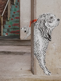 A mural of two black and white dogs is painted on the corner of a concrete wall. The larger dog appears to be in the foreground, with detailed line work illustrating its fur. The smaller dog is positioned behind it, partially hidden. A red stripe extends across the wall behind the dogs. In the background, there is a staircase with turquoise steps and red decorative patterns, leading up to a brick wall.