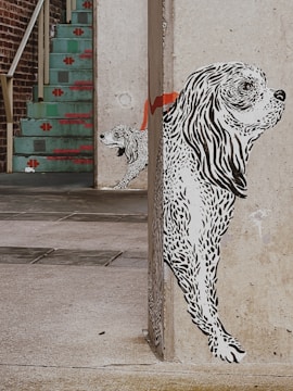 A mural of two black and white dogs is painted on the corner of a concrete wall. The larger dog appears to be in the foreground, with detailed line work illustrating its fur. The smaller dog is positioned behind it, partially hidden. A red stripe extends across the wall behind the dogs. In the background, there is a staircase with turquoise steps and red decorative patterns, leading up to a brick wall.