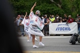 A cultural parade features two individuals in traditional attire, performing a dance on the street in front of a crowd. Their costumes are intricately designed with blue patterns and Japanese characters. The audience is diverse, with people taking photos and clapping behind a barrier. A large banner with the brand name 'ITOCHU' is prominently displayed.