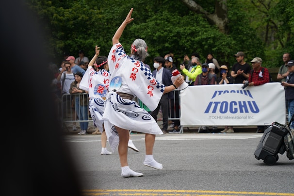 A cultural parade features two individuals in traditional attire, performing a dance on the street in front of a crowd. Their costumes are intricately designed with blue patterns and Japanese characters. The audience is diverse, with people taking photos and clapping behind a barrier. A large banner with the brand name 'ITOCHU' is prominently displayed.