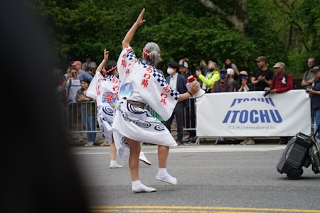 A cultural parade features two individuals in traditional attire, performing a dance on the street in front of a crowd. Their costumes are intricately designed with blue patterns and Japanese characters. The audience is diverse, with people taking photos and clapping behind a barrier. A large banner with the brand name 'ITOCHU' is prominently displayed.