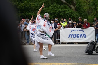 A cultural parade features two individuals in traditional attire, performing a dance on the street in front of a crowd. Their costumes are intricately designed with blue patterns and Japanese characters. The audience is diverse, with people taking photos and clapping behind a barrier. A large banner with the brand name 'ITOCHU' is prominently displayed.