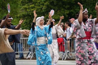 A group of people dressed in colorful traditional Japanese kimonos are performing a dance outdoors. The dancers are raising their arms with gestures, and some hold fans. The background features a crowd of onlookers behind metal barriers, suggesting a public event or festival. The setting is lush with greenery.