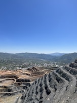 A large open-pit mine with terraced layers is visible in the foreground. Beyond the mine, a small town is nestled in a valley surrounded by lush green hills. The sky is clear and blue, suggesting a sunny day.