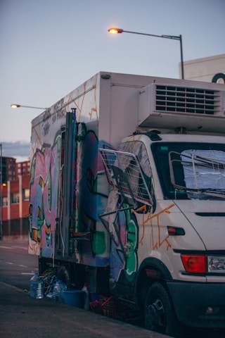 A truck is parked on the side of a street, covered in colorful graffiti art. The vehicle appears to be a repurposed food truck or similar vehicle with a large refrigeration unit on top. Several streetlights are illuminated as the sky transitions from day to night. Water bottles and other containers are visible on the ground next to the truck.