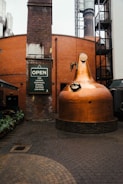 A large copper distillation apparatus is set in an industrial courtyard. The surrounding architecture features red brick walls and industrial elements, including a tall chimney and metal structures. A sign reads 'OPEN for laughs, cocktails, stories, tastings, and more.' The ground is paved with cobblestones and there are plants along the walkway.
