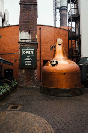 A large copper distillation apparatus is set in an industrial courtyard. The surrounding architecture features red brick walls and industrial elements, including a tall chimney and metal structures. A sign reads 'OPEN for laughs, cocktails, stories, tastings, and more.' The ground is paved with cobblestones and there are plants along the walkway.