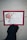 A hand wearing a black glove is holding a card featuring a decorative floral design with a large pink flower and green leaves. The card displays a religious text from John 3:16 in a black, elegant font. The border of the card is red with a thin green outline.