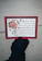 A hand wearing a black glove is holding a card featuring a decorative floral design with a large pink flower and green leaves. The card displays a religious text from John 3:16 in a black, elegant font. The border of the card is red with a thin green outline.