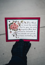 A hand wearing a black glove is holding a card featuring a decorative floral design with a large pink flower and green leaves. The card displays a religious text from John 3:16 in a black, elegant font. The border of the card is red with a thin green outline.