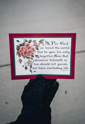 A hand wearing a black glove is holding a card featuring a decorative floral design with a large pink flower and green leaves. The card displays a religious text from John 3:16 in a black, elegant font. The border of the card is red with a thin green outline.