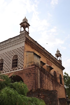 An old, weathered building featuring intricate architectural elements such as arches and domed turrets. The structure appears to be made of brick and stone with visible signs of age and decay. Vegetation is present in the foreground, and the sky is partly cloudy.