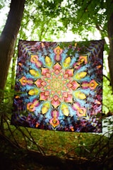 A vibrant tapestry with intricate, symmetrical, psychedelic patterns featuring kaleidoscopic designs. It is suspended between two trees in a lush, green forest setting, allowing sunlight to filter through the leaves.