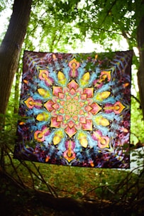 A vibrant tapestry with intricate, symmetrical, psychedelic patterns featuring kaleidoscopic designs. It is suspended between two trees in a lush, green forest setting, allowing sunlight to filter through the leaves.