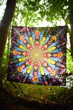 A vibrant tapestry with intricate, symmetrical, psychedelic patterns featuring kaleidoscopic designs. It is suspended between two trees in a lush, green forest setting, allowing sunlight to filter through the leaves.