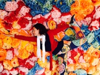 A person is performing a yoga pose, balancing against a vibrant backdrop of paper flowers in various colors including red, orange, yellow, blue, and green. They are wearing colorful, patterned leggings, a red sweater, and a white scarf.